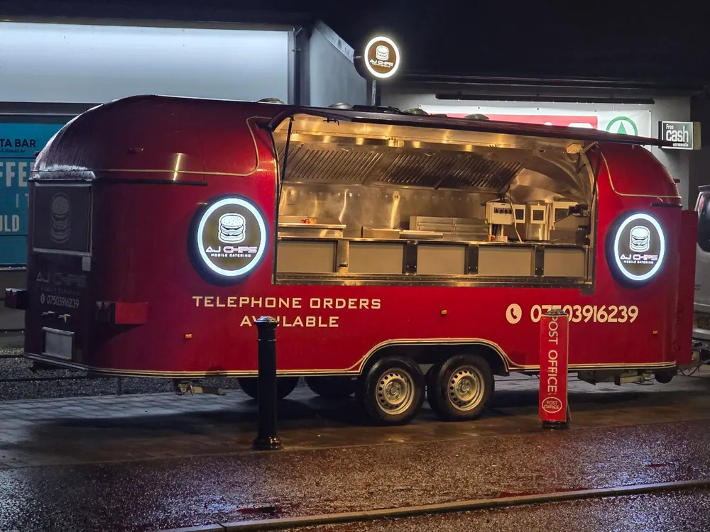 AJ Chips mobile catering trailer at night with illuminated circular logo lightboxes either side of the serving hatch.