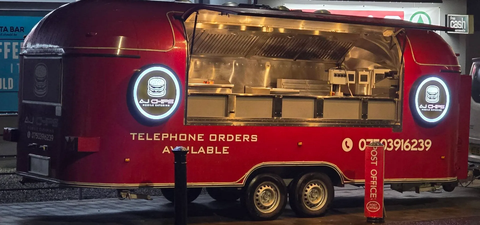 AJ Chips mobile catering trailer at night with illuminated circular logo lightboxes either side of the serving hatch.