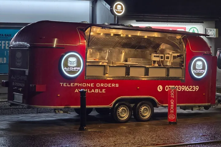AJ Chips mobile catering trailer at night with illuminated circular logo lightboxes either side of the serving hatch.