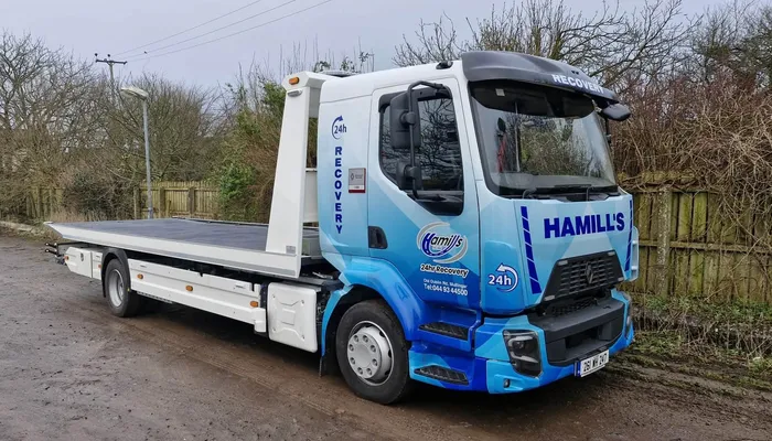 Hamill's 24hr Recovery flatbed truck in a blue geometric vinyl wrap, parked on a rural road.