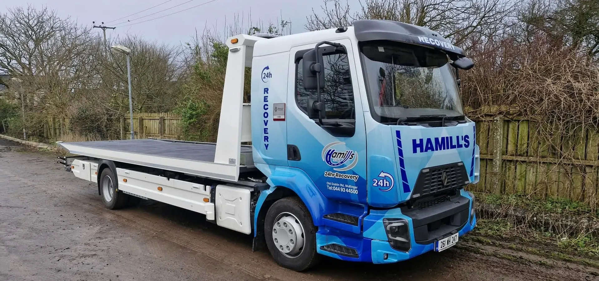 Hamill's 24hr Recovery flatbed truck in a blue geometric vinyl wrap, parked on a rural road.