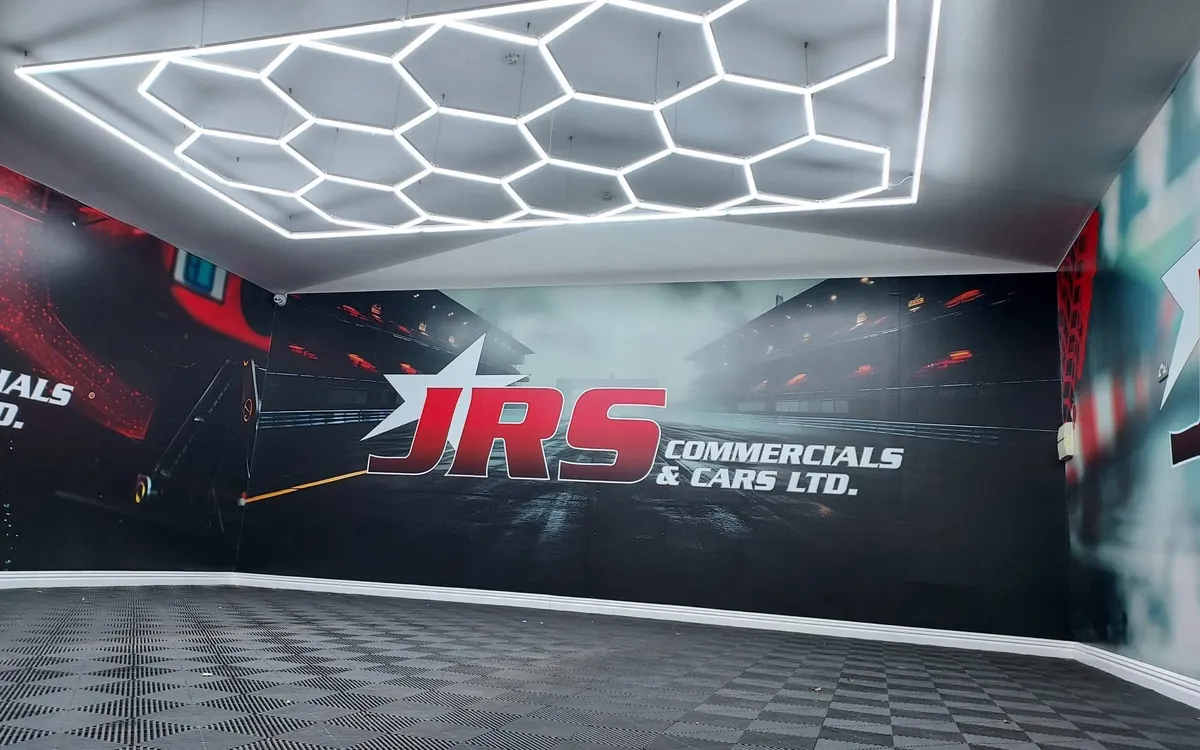JRS Commercials & Cars showroom wall graphic with a wet racetrack scene and the JRS logo, under hexagonal LED lighting.