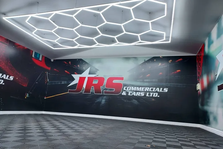 JRS Commercials & Cars showroom wall graphic with a wet racetrack scene and the JRS logo, under hexagonal LED lighting.