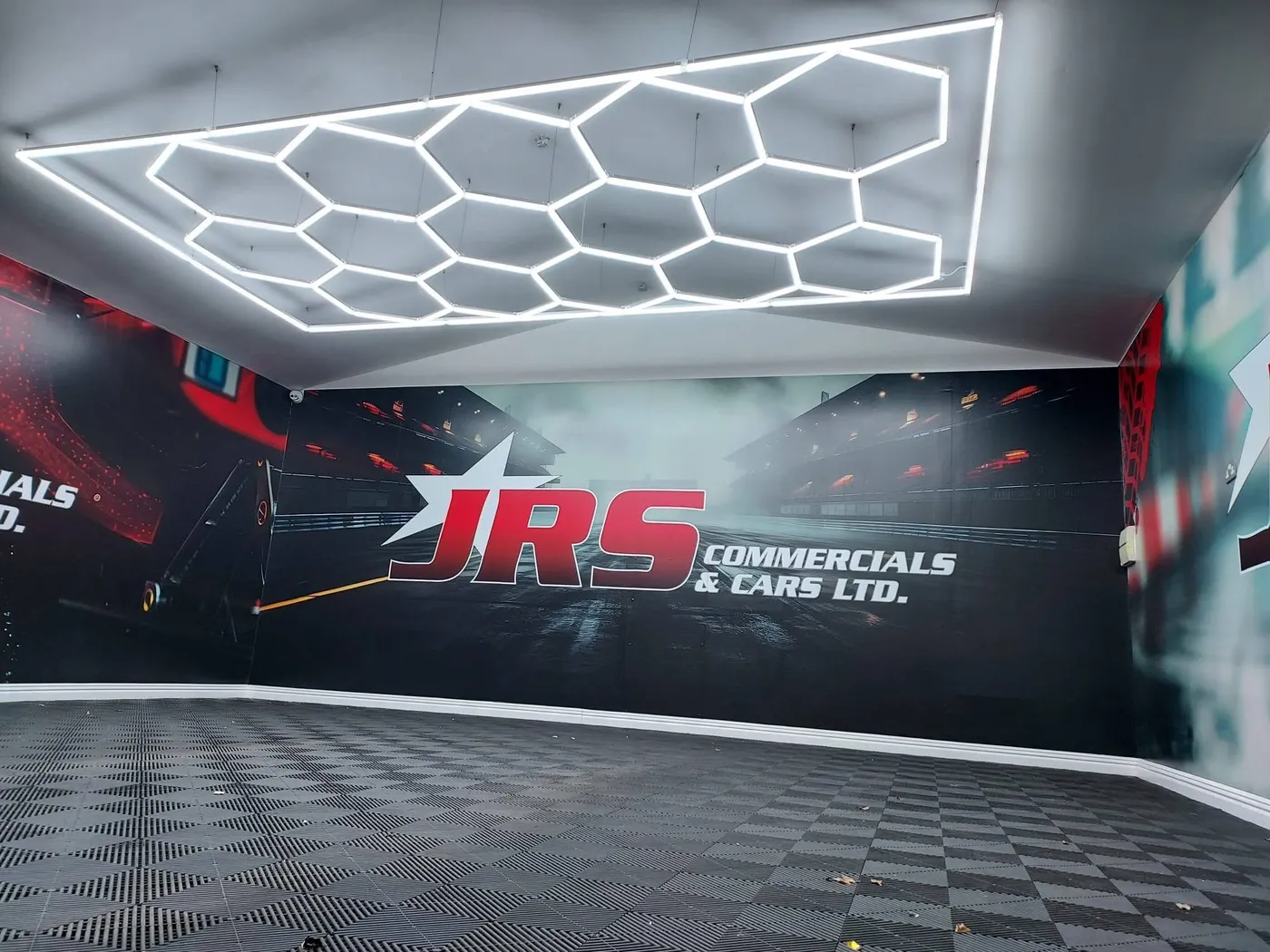 JRS Commercials & Cars showroom wall graphic with a wet racetrack scene and the JRS logo, under hexagonal LED lighting.