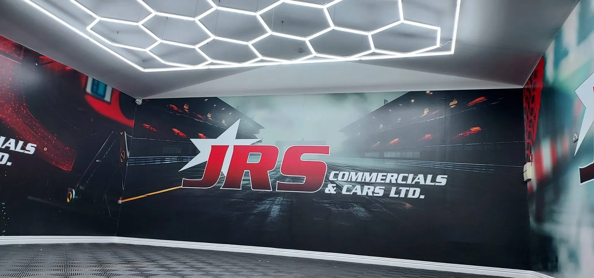 JRS Commercials & Cars showroom wall graphic with a wet racetrack scene and the JRS logo, under hexagonal LED lighting.