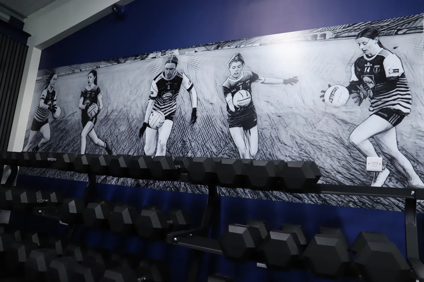 Scotstown GAA gym feature wall showing a black-and-white action sequence of players above a weights rack.