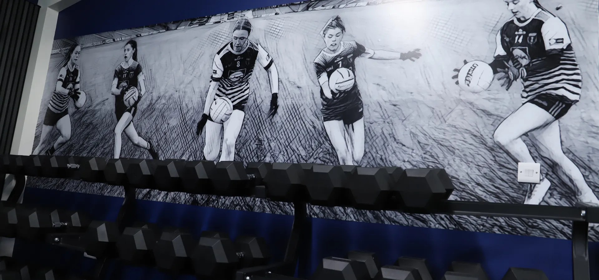 Scotstown GAA gym feature wall showing a black-and-white action sequence of players above a weights rack.