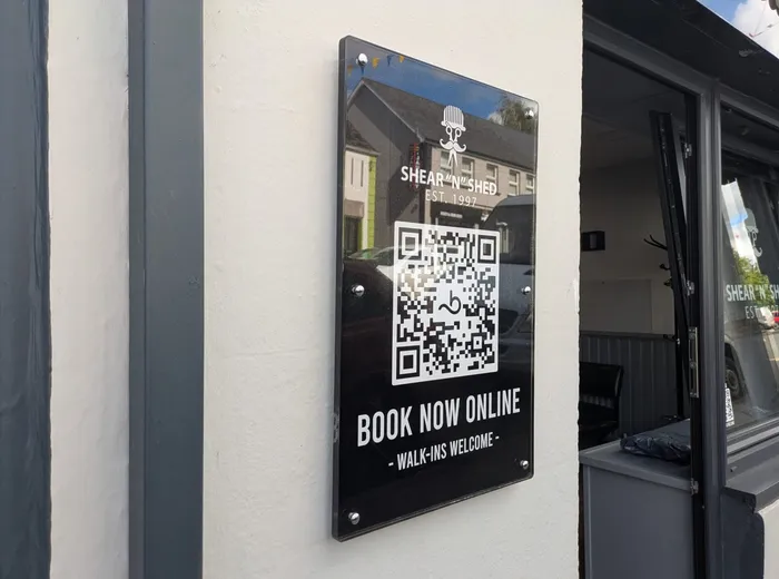 Shear 'n' Shed black acrylic plaque with QR code and Book Now Online, mounted on a white wall with silver standoff fixings.